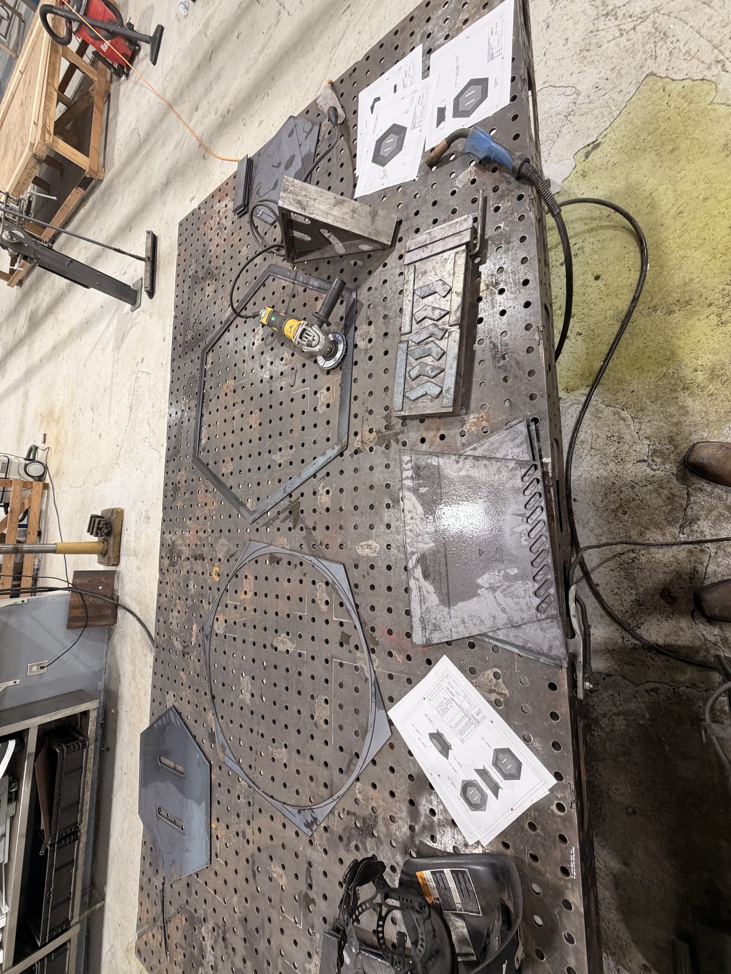 Plasma-cut fire pit parts on the weld table: a hex ring, a round ring, slotted panels, and hex plates, with drawings beside them. The Primo logo is in the lower right